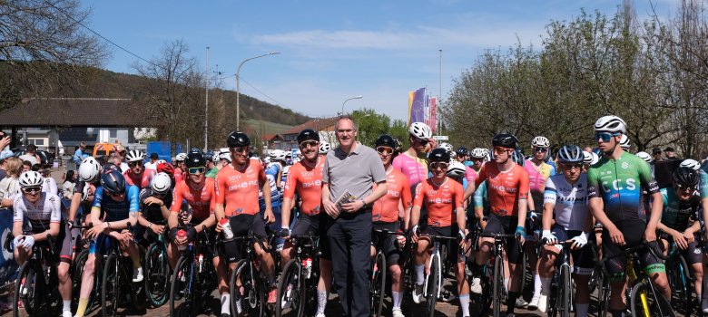 Landrat Dietmar Seefeldt steht kurz vor dem Start des Großen Preises der Südlichen Weinstraße inmitten des Teams Bike Aid Südliche Weinstrasse. Neben den Bike Aid Fahrern in ihren orangen Trikots stehen auch bereits andere Teilnehmer mit ihren Rädern am Start.