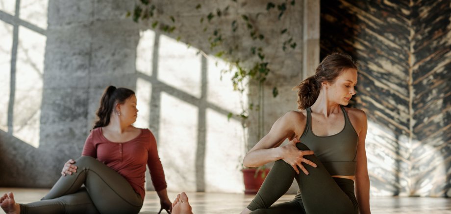 Zwei Frauen sitzen auf Yogamatten und machen Dehnungsübungen. 