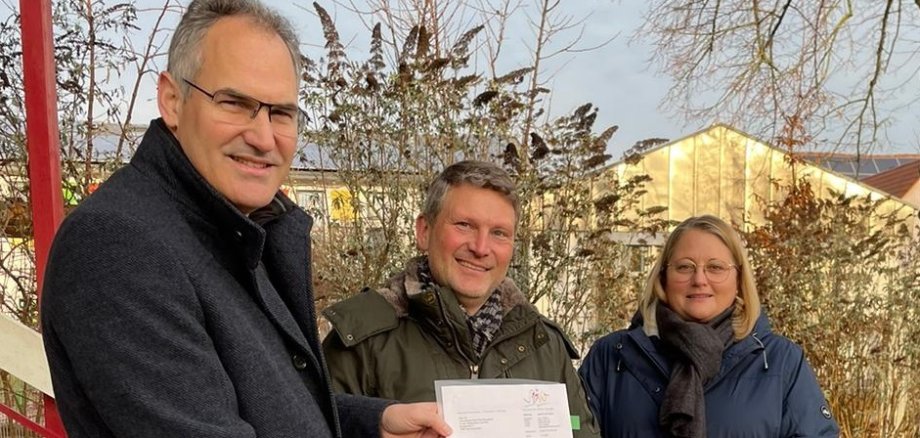 Landrat Dietmar Seefeldt (links) übergibt Ortsbürgermeister Matthias Neufeld und Beigeordneter Diana Nowak den Förderbescheid für die kommunale Kita.