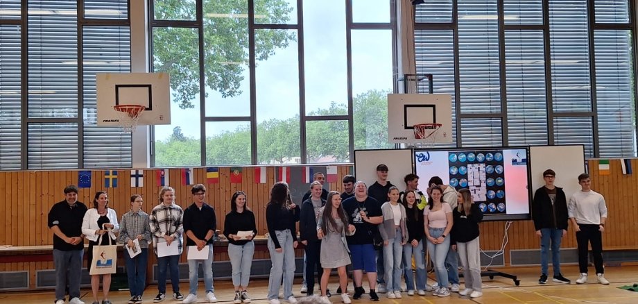 Schülerinnen und Schüler stehen in einer Turnhalle, dahinter verschiedene Nationalflaggen.