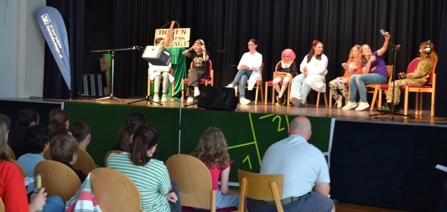 Eine Bühne mit acht jungen Menschen in verschiedenen Kostümen, die auf Stühlen sitzen und an einer Theatervorführung oder einem Bühnenprogramm teilnehmen. Im Vordergrund des Bildes sitzt ein Publikum, das aus Kindern und Erwachsenen besteht.