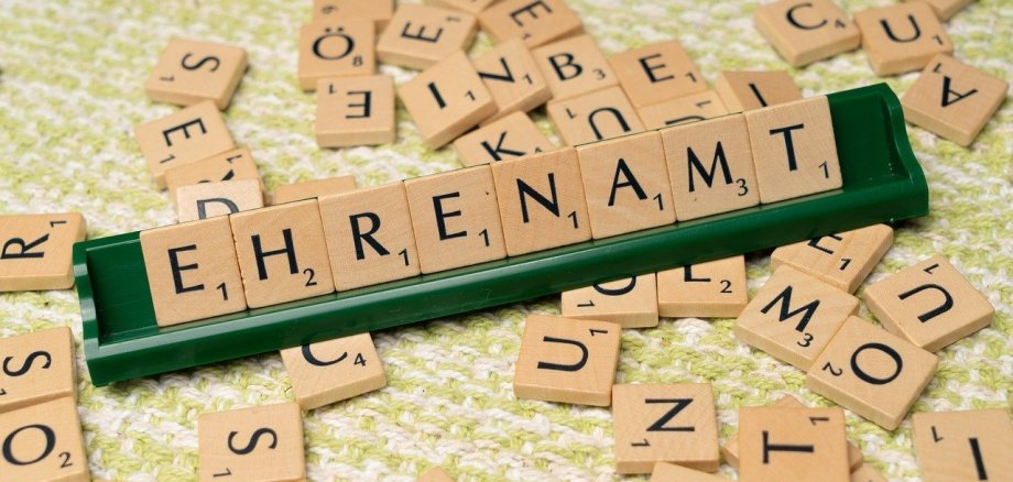 Scrabble-Buchstaben, die das Wort 'EHRENAMT' bilden, liegen auf einem grünen Buchstabenhalter. Weitere verstreute Scrabble-Buchstaben sind im Hintergrund auf einem grünen und weißen Teppich zu sehen.