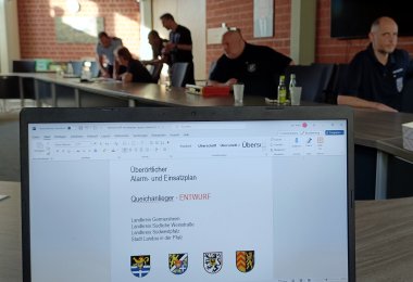 Im Vordergrund ein Bildschirm, auf dem ein Dokument geöffnet ist mit Titel "Überörtlicher Alarm- und Einsatzplan", im Hintergrund sitzen Männer des Katastrophenschutzes an Tischen.