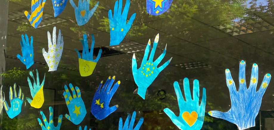 Mehrere ausgeschnittene Handabdrücke in Blau- und Gelbtönen sind an einem Fenster angebracht. Einige der Handabdrücke sind mit Sternen und Herzen verziert, andere haben Streifen oder Punkte. Die Gestaltung der Handabdrücke zeigt kreative und farbenfrohe Muster. Durch das Fenster ist der Innenraum eines Klassenzimmers mit Tischen und Stühlen zu sehen.