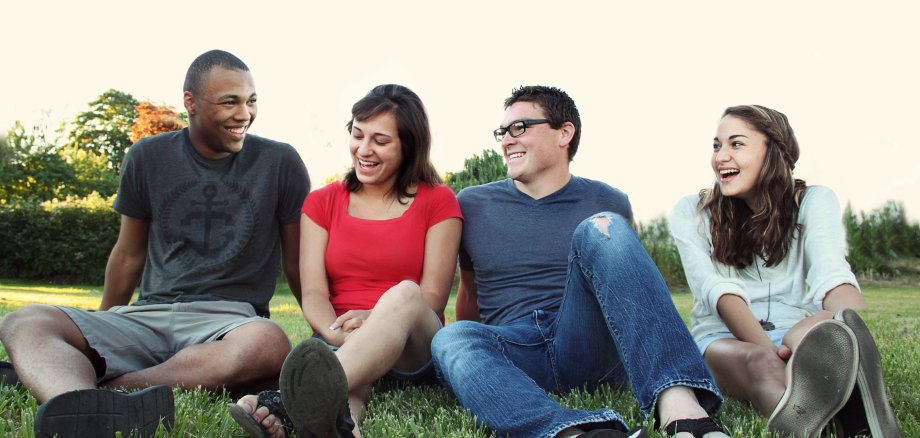 Eine Gruppe von vier jungen Erwachsenen sitzt lachend auf dem Gras. Von links nach rechts tragen sie: ein schwarzes T-Shirt und graue Shorts, ein rotes T-Shirt und schwarze Sandalen, ein dunkelblaues T-Shirt und Jeans mit einer Brille, sowie ein weißes Hoodie und Shorts. Im Hintergrund sind Bäume und ein heller Himmel zu sehen.