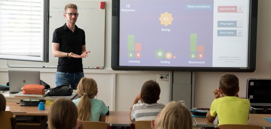 Ein junger Lehrer steht vor einer Klasse und unterrichtet mit Hilfe eines Smartboards.  Die jungen Schülerinnen und Schüler hören aufmerksam zu.