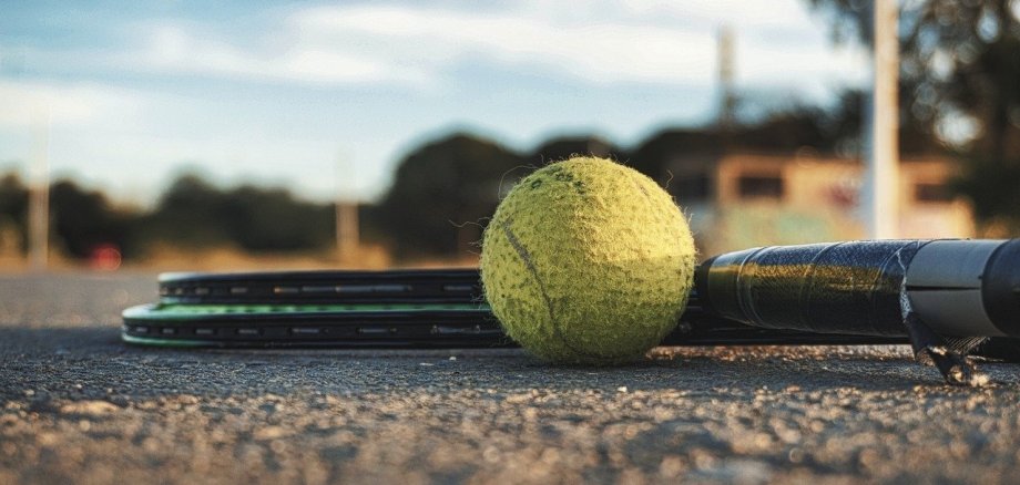 Nahaufnahme eines Tennisballs und zweier Tennisschläger, die auf dem Boden eines Tennisplatzes liegen. Der Hintergrund ist verschwommen.