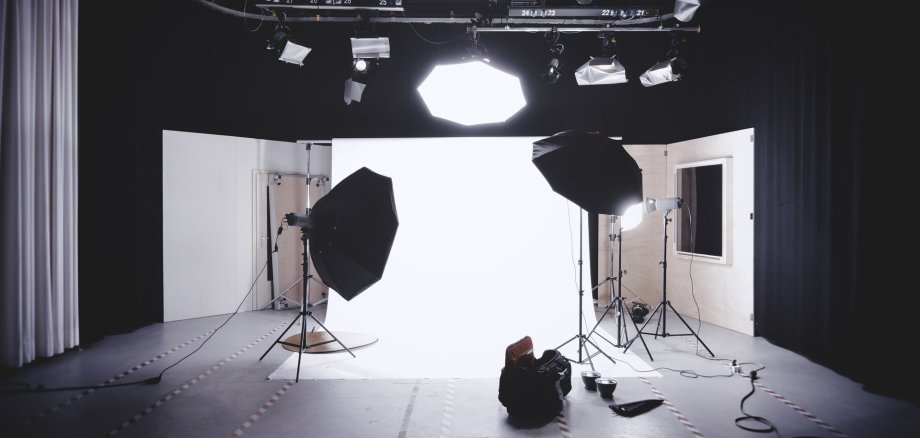 Professionelles Fotostudio mit Beleuchtungs-Setup und weißen Hintergrundwänden. Mehrere Studioleuchten und Softboxen sind um eine leere Fläche herum positioniert, bereit für ein Fotoshooting. Im Vordergrund befinden sich Kabel und eine Tasche mit Ausrüstung, während im Hintergrund große Softboxen und Lichtquellen an der Decke befestigt sind (generiert mit Hilfe von KI).