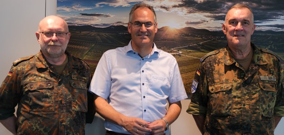 Landrat Dietmar Seefeldt empfängt die Oberstleutnante der Bundeswehr Kay Wittenborn und Wolfgang Eder im Kreishaus. Auf dem Bild stehen (von links nach rechts) die zwei uniformierten Oberstleutnante der Bundeswehr neben Landrat Seefeldt, der ein helles Hemd trägt. Im Hintergrund ist eine malerische Landschaft mit Feldern und einem Sonnenuntergang zu sehen, die auf einem Wandbild abgebildet ist. Die beiden Bundeswehrangehörigen tragen typische Bundeswehr-Tarnuniformen und die deutschen Flaggenabzeichen auf ihren Ärmeln.