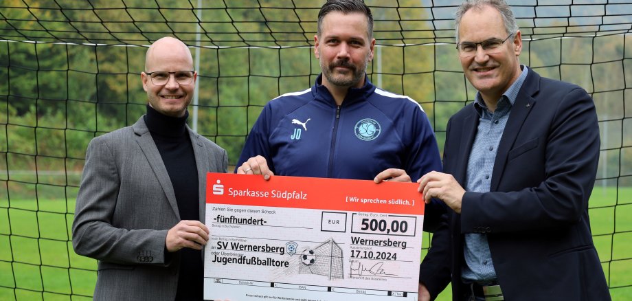 Sparkassenvorsitzender Svend Larsen, Jan Ohlert vom SV Wernersberg und Landrat Dietmar Seefeldt stehen vor einem Fußballtor und halten einen großen symbolischen Scheck der Sparkasse Südpfalz über 500 Euro in die Kamera. Der Scheck ist an den SV Wernersberg für die Unterstützung der Jugendarbeit im Fußballverein adressiert. Hintergrund zeigt ein grünes Fußballfeld und eine hügelige Landschaft.