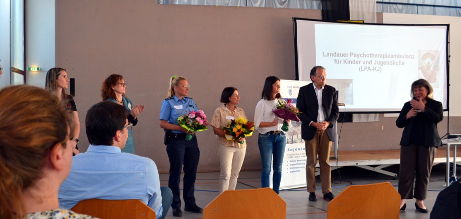 Auf dem Bild stehen mehrere Personen in einer Halle auf einer Bühne. Drei Frauen, darunter eine Polizistin, halten Blumensträuße in den Händen und werden von den umstehenden Personen, darunter auch ein Mann im Anzug, applaudiert. Im Hintergrund ist eine Leinwand zu sehen, auf der die „Landauer Psychotherapieambulanz für Kinder und Jugendliche (LPA-KJ)“ vorgestellt wird. Vor der Bühne sitzen weitere Personen und folgen der Veranstaltung.