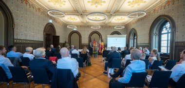 Adrian Rinck und Prof. Dr. Matthias Bahr präsentieren das Projekt „Youth. Europe. Music.“ während der Preisverleihung im historischen Festsaal in Weimar, in dem das politische Austauschformat des „Weimarer Dreiecks“ vor 33 Jahren gegründet wurde. Im Vordergrund sitzen zahlreiche Zuschauer der Preisverleihung auf Stühlen.