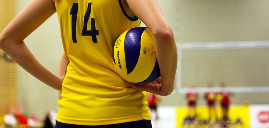 Oberkörper einer Frau im Trikot im Vordergrund, sie trägt einen Volleyball. Im Hintergrund Volleyballnetz und weiter Sportlerinnen.