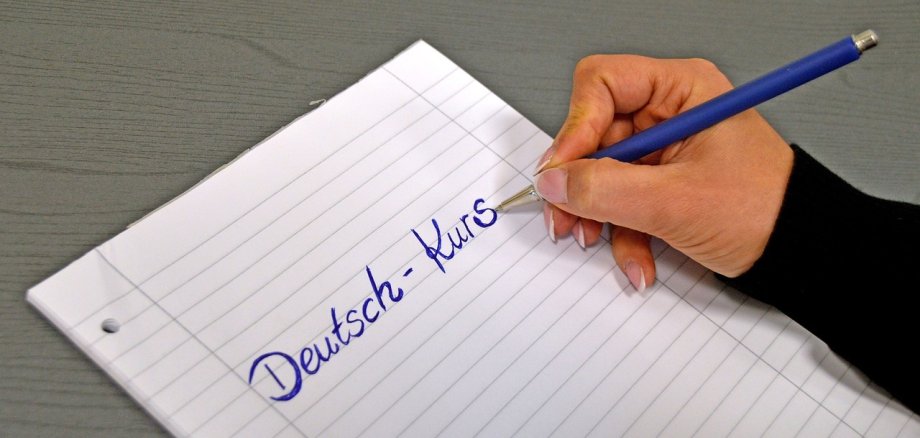Eine Frau schreibt mit Kugelschreiber den Begriff "Deutsch-Kurs" auf ein Blatt Papier.